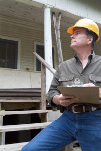 An inspector in a hardhat holds a clipboard as he evaluates a home exterior.